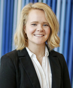 woman with shoulder-length blond hair, wearing a white shirt and black blazer stands against a background with multiple shades of blues.