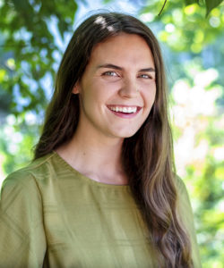 A photo of a girl with ling brown hair wearing a green shirt standing outside and smiling